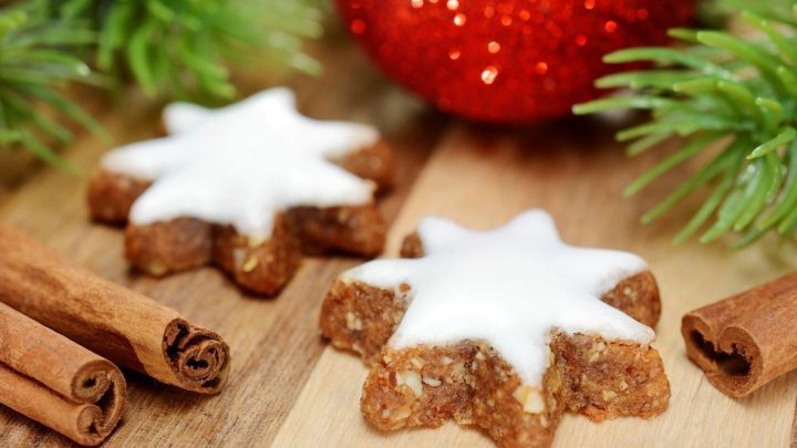 Zwei weihnachtliche Pl&auml;tzchen mit Zuckerguss liegen auf einem Holzbrett, umgeben von Zimtstangen und einer roten Dekorkugel.