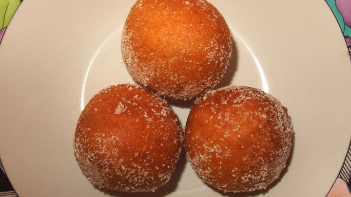 Drei goldbraun gebratene Quarkb&auml;llchen liegen auf einem wei&szlig;en Teller, umgeben von buntem Geschirr.