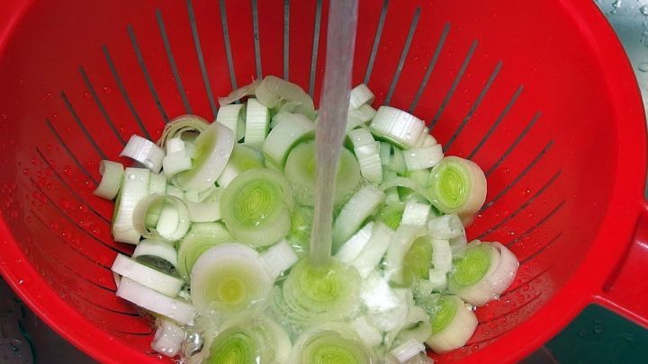 In einem roten Sieb werden geschnittene, frische Lauchst&uuml;cke mit Wasser abgesp&uuml;lt, um sie f&uuml;r die Zubereitung von gesundem, gr&uuml;nem Gem&uuml;se vorzubereiten.