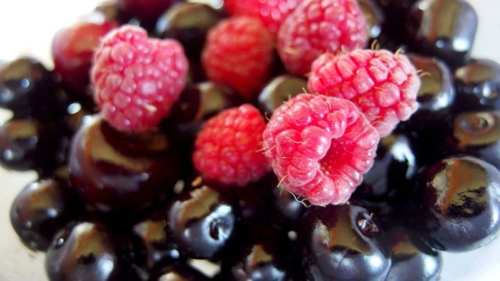 Himbeeren und dunkle Beeren liegen auf einem wei&szlig;en Teller und gl&auml;nzen im Licht.