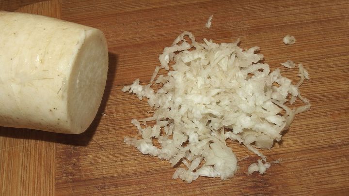 Eine frisch geriebene Meerrettich-H&auml;ufchen liegt neben einer ganzen Meerrettichwurzel auf einem Holzbrett.