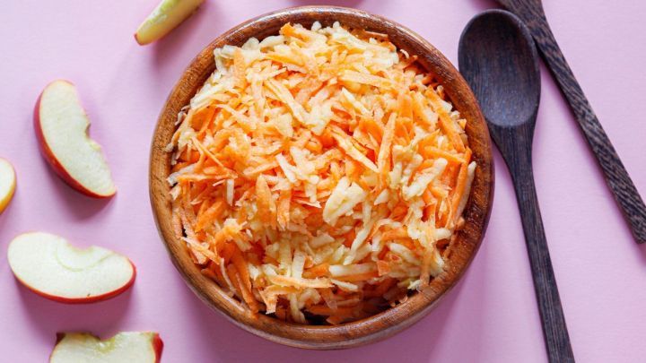 Rohkostsalat aus geriebenen &Auml;pfeln und M&ouml;hren liegt in einer Sch&uuml;ssel, umgeben von Apfelscheiben auf rosa Hintergrund.
