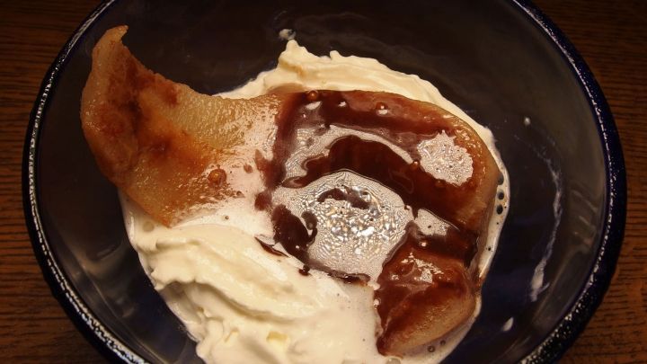 Eine angeregte Birne liegt in einer Sch&uuml;ssel mit cremiger Schokoladensauce und Sahne, umgeben von einem dunklen Holzuntergrund.