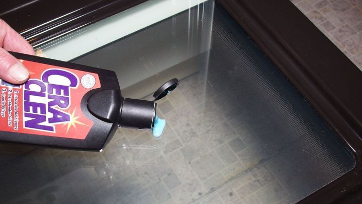 Eine Hand h&auml;lt eine Flasche Glaskeramikreiniger &uuml;ber ein Backofensichtfenster und bereitet das Reinigen vor.