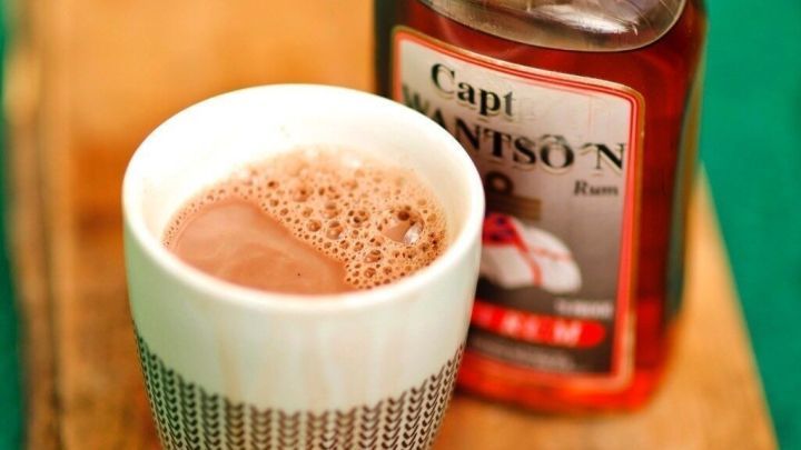 Ein elegantes Glas mit hei&szlig;em Kakao und Rum steht auf einem Holzbrett neben einer Flasche Rum, umgeben von einer einfachen gr&uuml;nen Umgebung.