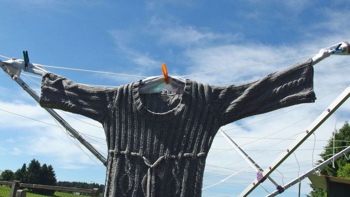 Ein grauer Pullover h&auml;ngt zum schonenden Trocknen auf einer W&auml;scheleine unter blauen Himmel und gr&uuml;nen B&auml;umen.
