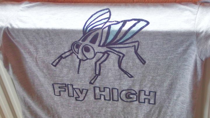Ein T-Shirt mit einem gro&szlig;en, cartoonhaften Fliegenmotiv und dem Text &bdquo;Fly HIGH&ldquo; h&auml;ngt an einer W&auml;scheleine.