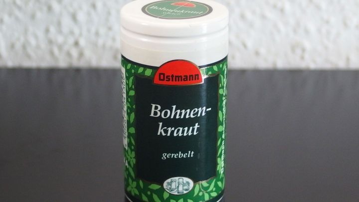 Ein Beh&auml;lter mit Bohnenkraut steht auf einem dunklen Tisch, w&auml;hrend der Hintergrund eine helle Wand zeigt.