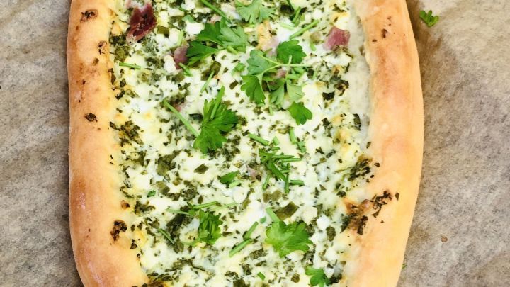 Ein ovales Fladenbrot liegt auf Backpapier, belegt mit einer cremigen Mischung und frischen Kr&auml;utern.