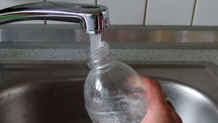 Eine PET-Flasche wird unter einem Wasserhahn gef&uuml;llt, w&auml;hrend die Hand sie h&auml;lt, in einer K&uuml;che mit Fliesen.