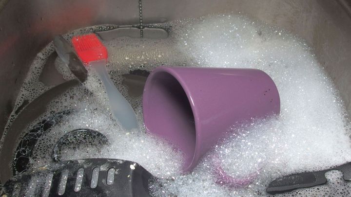 Ein lila Becher liegt schr&auml;g in einer Sp&uuml;lmaschine, umgeben von Schaum und Geschirr, w&auml;hrend die Klappe der Maschine offen ist.