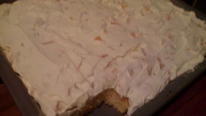 Ein gro&szlig;er Fantakuchen mit Pfirsichen und Sahne&uuml;berzug steht auf einer Holzplatte in einer gem&uuml;tlichen K&uuml;che.