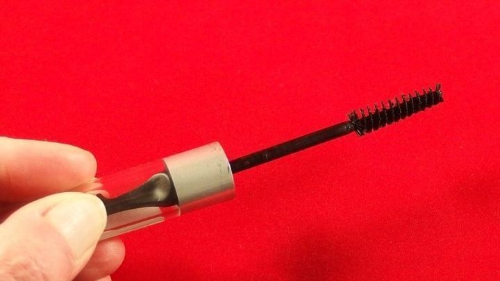 Eine Hand h&auml;lt eine Wimperntusche hoch, w&auml;hrend sie vor einem roten Hintergrund gezeigt wird, was auf eingetrocknete Wimperntusche hinweist.