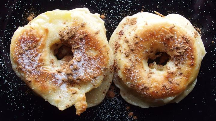 Frittierte Apfelk&uuml;chle liegen goldbraun auf einer dunklen Oberfl&auml;che, bestreut mit etwas Zucker, bereit zum Genie&szlig;en.