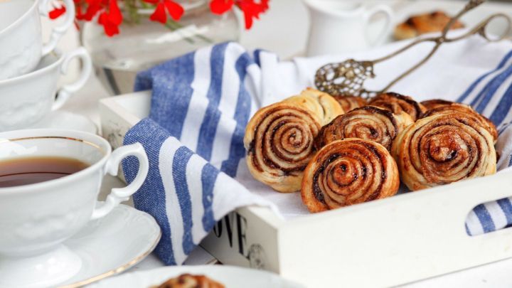 Frisch gebackene Nutella-Schnecken liegen in einem wei&szlig;en Korb auf einem blauen Tuch, umgeben von Teetassen und Blumen.