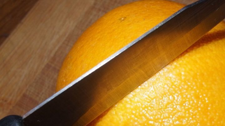 Eine orangefarbene Apfelsine liegt auf einem Holzbrett, w&auml;hrend ein gl&auml;nzendes Messer darauf zum Sch&auml;len positioniert ist.