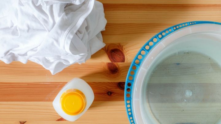 Wei&szlig;es T-Shirt liegt neben einer Sch&uuml;ssel mit Wasser und einer Flasche Essigreiniger auf einem Holztisch.
