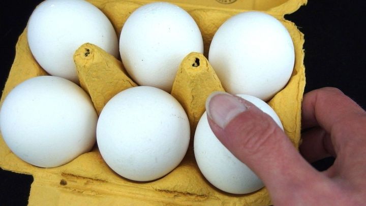 Sechs wei&szlig;e Eier liegen in einer gelben Schachtel, w&auml;hrend eine Hand ein Ei herausnimmt, um ein kaputtes zu vermeiden.