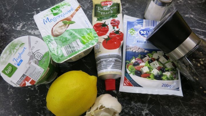 Zutaten liegen auf einer schwarzen Marmorfl&auml;che: Joghurt, Magerquark, Tomatenmark, eine Zitrone, Knoblauch, Feta, Salz, Pfeffer.