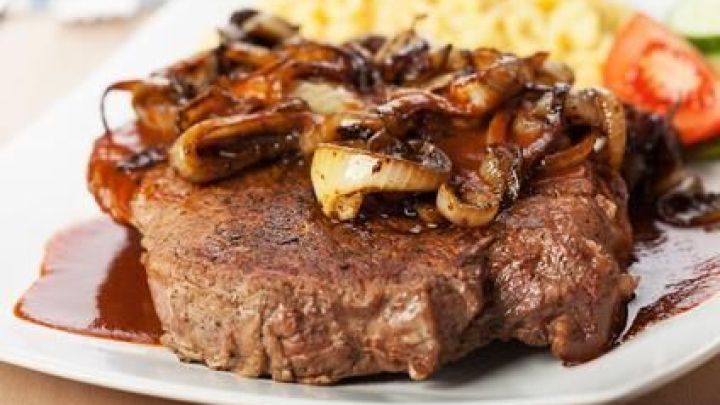 Zwiebelrostbraten liegt auf einem Teller, bedeckt mit gebratenen Zwiebeln und So&szlig;e, daneben Sp&auml;tzle und Gem&uuml;se.