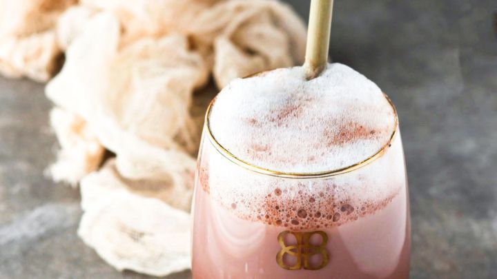 Ein Glas mit selbstgemachtem Baileys steht auf einer grauen Oberfl&auml;che, umgeben von einem Tuch und einem Eisw&uuml;rfel.