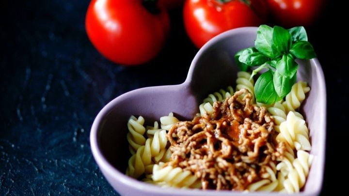 Eine lila herzf&ouml;rmige Sch&uuml;ssel enth&auml;lt Nudeln mit Bolognese-Sauce, dekoriert mit frischem Basilikum, umgeben von roten Tomaten.