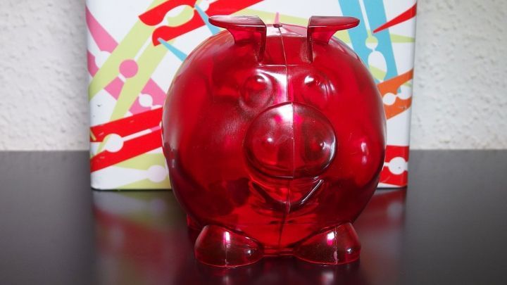 Ein rotes Sparschwein steht auf einem Tisch und wartet darauf, mit Waschmittelkasse gef&uuml;llt zu werden. Hintergrund bunt.