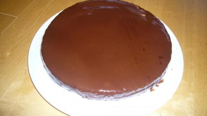 Schokoladentorte steht mit gl&auml;nzender Glasur auf einem wei&szlig;en Teller, arrangiert auf einem h&ouml;lzernen Tisch.