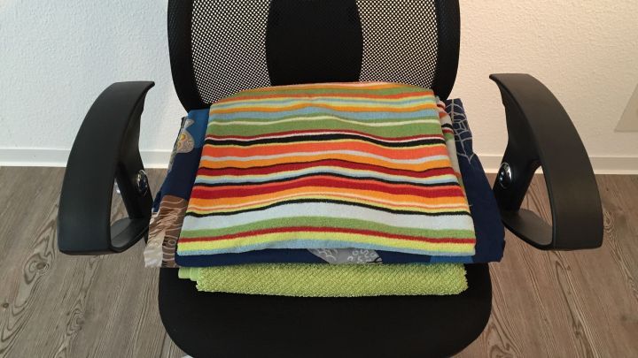 Ein Stapel mit bunt gestreiften Handt&uuml;chern liegt ordentlich auf einem schwarzen B&uuml;rostuhl in einem hellen Raum.