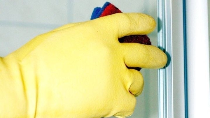 Eine Hand mit einem gelben Gummihandschuh reinigt das Duscht&uuml;rglas mit einem Schwamm, um Seifenreste zu entfernen.