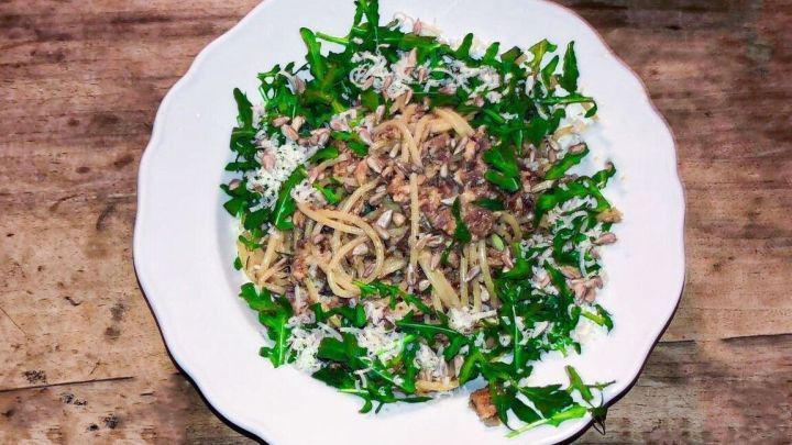 Ein Teller Nudeln mit frischen Kr&auml;utern und einer fleischigen So&szlig;e steht auf einem Holztisch und wirkt appetitlich.