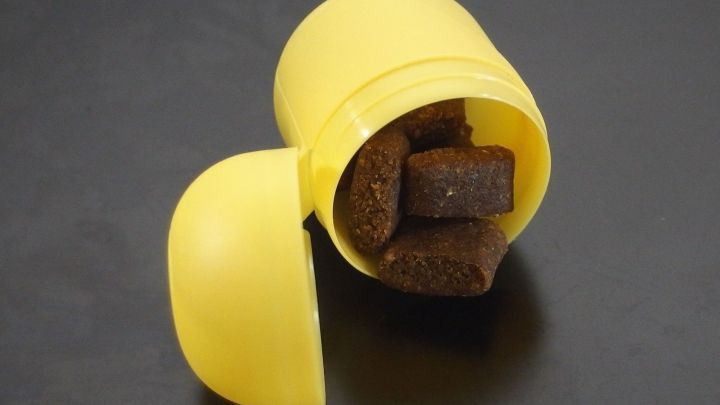 Eine gelbe &Uuml;-Ei-H&uuml;lle liegt ge&ouml;ffnet da, mit braunen Leckerlis f&uuml;r Hunde sichtbar darin, auf schwarzem Untergrund.