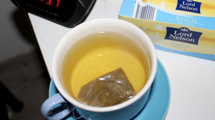 Eine Tasse mit Kamillentee steht auf einem Nachttisch. Der Teebeutel zieht in der warmen Fl&uuml;ssigkeit, w&auml;hrend die Uhr 21:51 anzeigt.