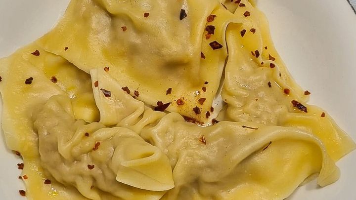 Auf einem wei&szlig;en Teller liegen dampfende Maultaschen, die mit roten Chiliflocken bestreut sind, auf einem Holztisch.