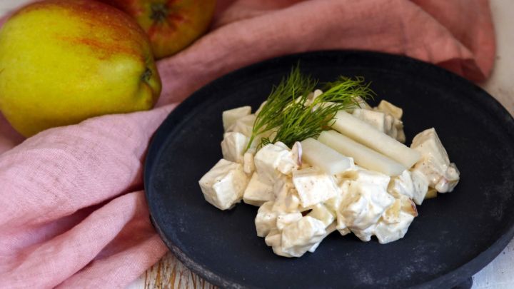 Kohlrabisalat liegt auf einem schwarzen Teller, umgeben von &Auml;pfeln und einem rosa Tuch, frische Kr&auml;uter garnieren das Gericht.