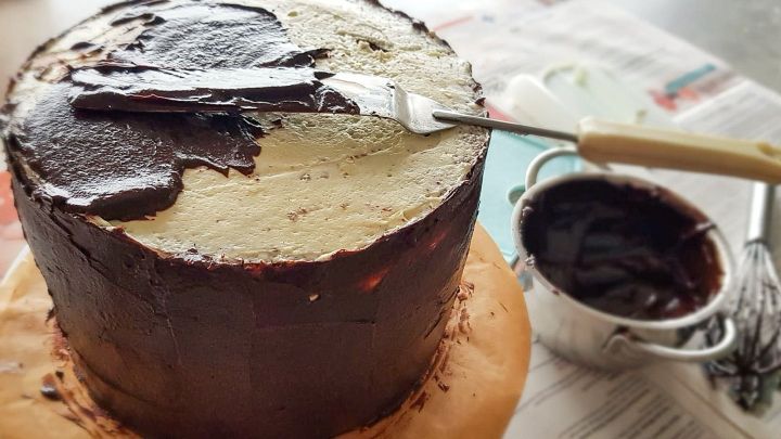 Ein Schokoladenkuchen wird mit einem Spachtel glatter gestaltet, w&auml;hrend er auf einem Tisch mit Zeitungen steht.