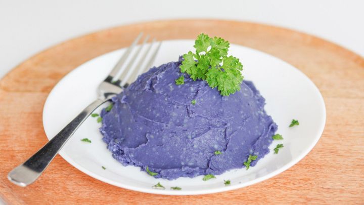 Violettes Kartoffelp&uuml;ree mit frischer Petersilie auf einem wei&szlig;en Teller wird auf einem Holzuntersetzer pr&auml;sentiert.
