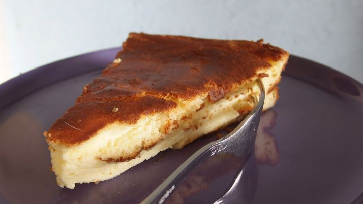 Ein St&uuml;ck K&auml;sekuchen ohne Boden liegt auf einem lila Teller, w&auml;hrend eine Gabel daneben bereit ist, um zu genie&szlig;en.