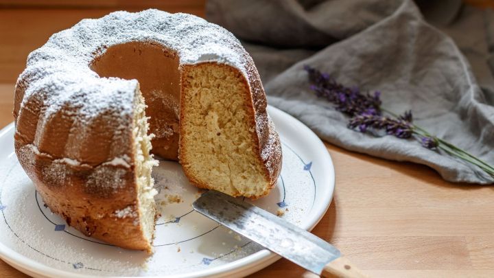 Ein Amaretto-Gugelhupf steht auf einem Teller, leicht angeschnitten, umgeben von einem grauen Tuch und einer Holzoberfl&auml;che.