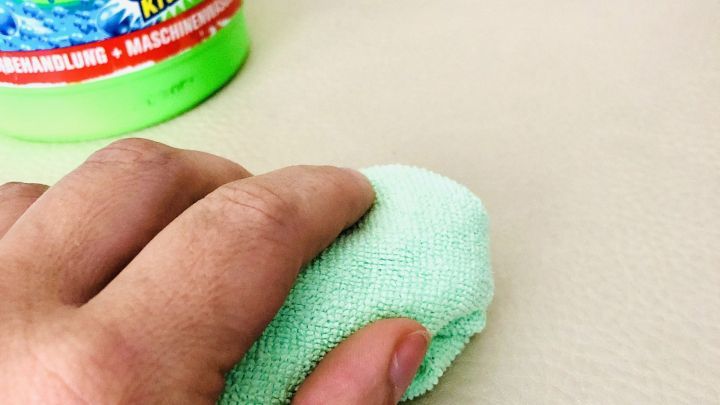 Ein Handtuch reinigt ein helles Kunstledersofa in einer hellen Umgebung mit einem Reinigungsmittel im Hintergrund.