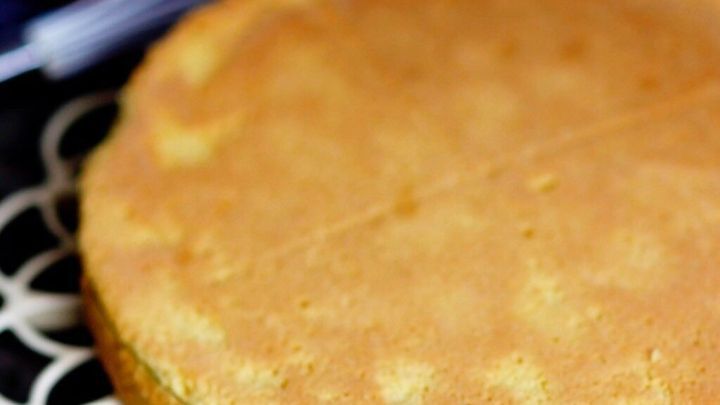 Ein bisquitfarbener Tortenboden liegt auf einer dekorativen Unterlage, umgeben von Eiern und einem Schneebesen.