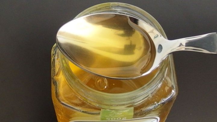 Ein L&ouml;ffel mit gl&auml;nzendem Honig schwebt &uuml;ber einem offenen Honigglas auf einem dunklen Hintergrund.