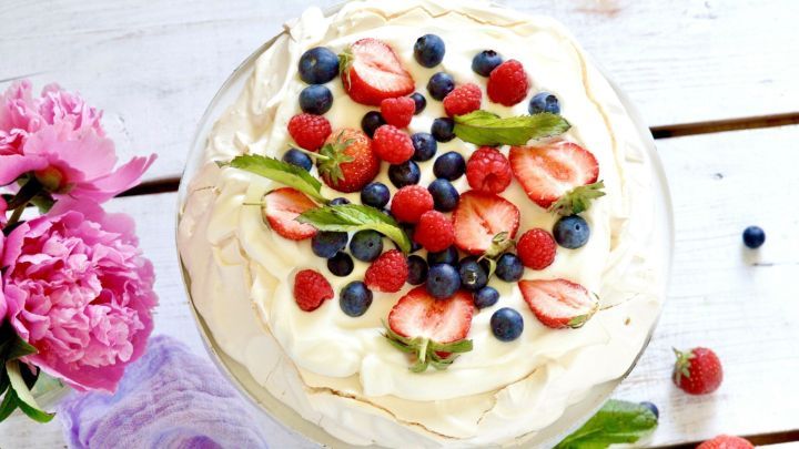 Eine farbenfrohe Beerentorte mit Baisers, mit frischen Beeren und Minze dekoriert, steht auf einem Holztablett.