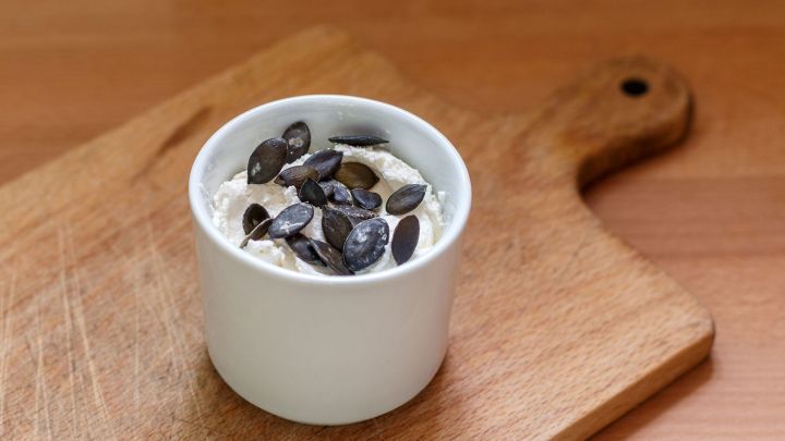 Ein kleiner, wei&szlig;er Becher mit K&uuml;rbiskernaufstrich steht auf einem Holzbrett, dekoriert mit K&uuml;rbiskernen.