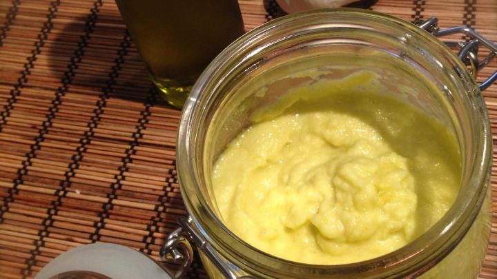 Ein Glas mit gelblichem Knoblauchp&uuml;ree steht auf einem Holzuntergrund, daneben eine Flasche &Ouml;l und eine Knoblauchknolle.