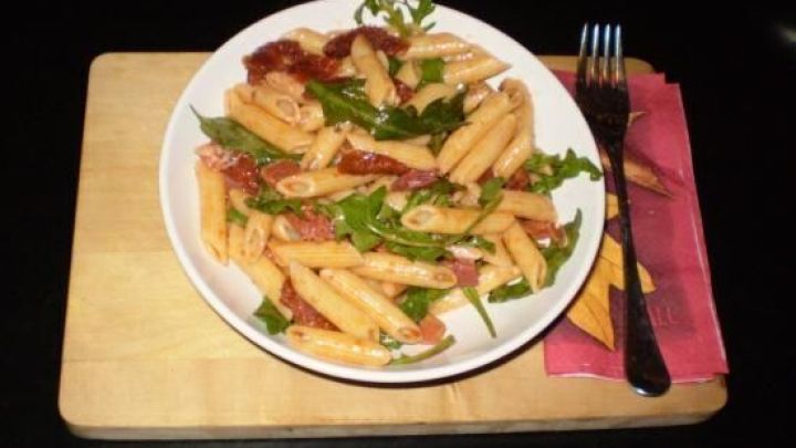 Ein Teller Nudelsalat Mediterrana liegt auf einem Holzbrett, umgeben von einem Besteck, bereit zum Genie&szlig;en.