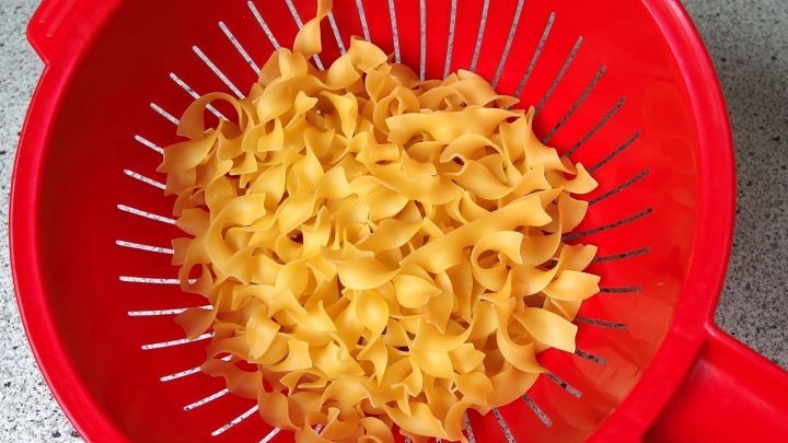 Gelbe Nudeln liegen in einem roten Sieb, w&auml;hrend sie im K&uuml;chenschaum abtropfen und auf den n&auml;chsten Kochschritt warten.