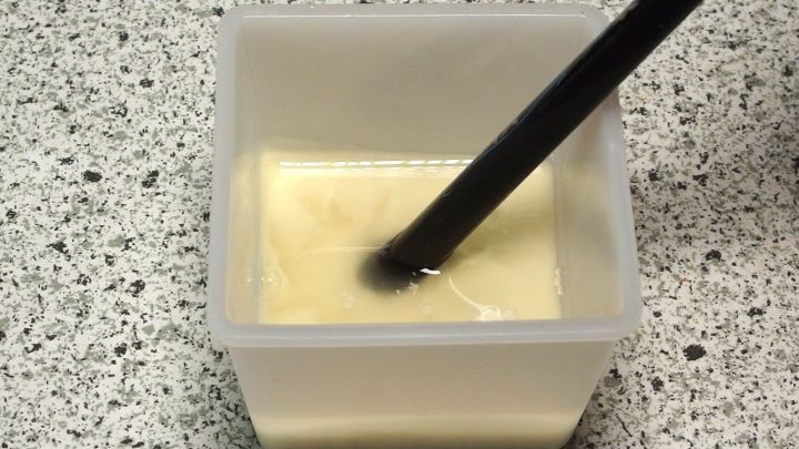 Ein schwarzer Pinsel taucht in eine schaumige, gelbe L&ouml;sung in einem transparenten Beh&auml;lter auf einer K&uuml;chenarbeitsfl&auml;che.