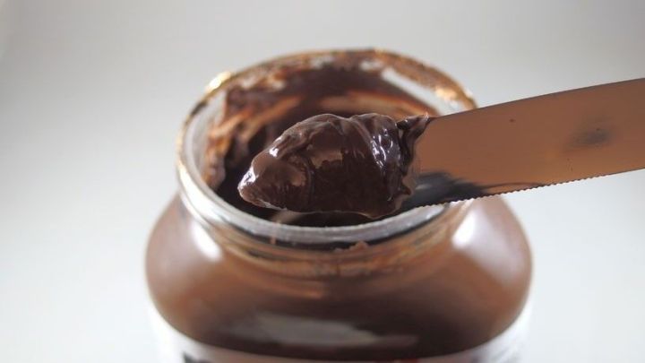 Ein Essl&ouml;ffel schokoladiger Nutella wird aus einem Glas entnommen, das auf einem hellen Untergrund steht.