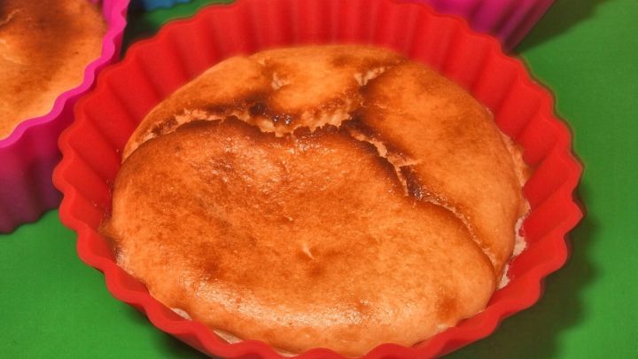 K&auml;semuffins stehen in bunten Silikonformen auf einer gr&uuml;nen Unterlage und zeigen eine goldbraune, leicht aufgerissene Oberfl&auml;che.